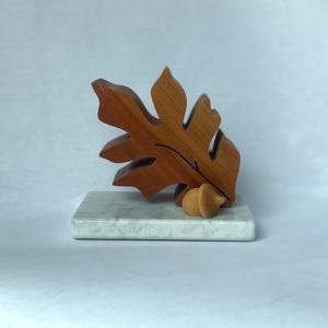 May include: A carved wooden oak leaf sculpture with a smooth, reddish-brown finish, featuring a single carved wooden acorn at its base. The entire piece is mounted on a rectangular, white marble base with subtle grey veining throughout.