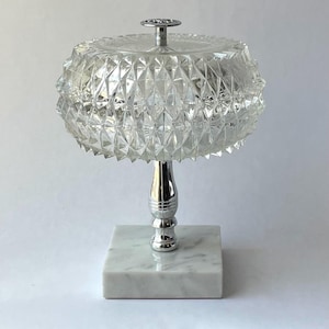 May include: A clear glass decorative object with a diamond-cut pattern, topped with a silver handle. It sits on a silver stem and a square white marble base. The object is likely a decorative piece or a container.