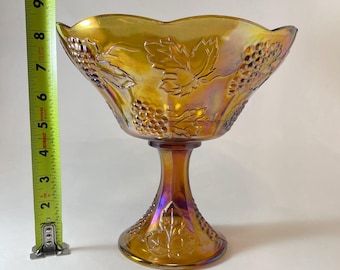 Pode incluir: Uma tigela vintage de vidro âmbar com base de pedestal. A tigela é decorada com um desenho de videira e tem um acabamento iridescente.