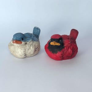 May include: Two ceramic bird figurines. One is a bluebird with a white chest and orange beak. The other is a red cardinal with a black face and yellow beak. Both birds are perched and facing forward.
