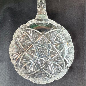May include: Clear, cut-glass candy dish with a handle. The dish features a starburst and geometric pattern. The rim has a scalloped edge. The handle is rectangular and has a decorative design. The dish is likely vintage.