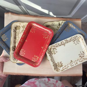 May include: A collection of rectangular metal trays featuring rounded corners and raised edges. The trays are finished in solid red, cream, and blue colors, each decorated with a repeating gold floral pattern along the borders and corners.