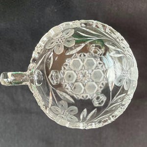 May include: Clear glass serving bowl with a handle, featuring floral and geometric etched designs. The bowl's rim and handle are intricately detailed, showcasing a vintage aesthetic. The bowl is likely used for serving snacks or desserts.