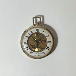 May include: A gold-tone pocket watch with a white dial featuring black Roman numerals. The center of the watch is skeletonized, revealing the internal gold-colored mechanical movement. The text Ever Swiss and Swiss Made is printed on the dial.