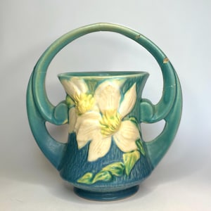 May include: A teal ceramic vase with a large handle. The vase features a floral design with white and yellow flowers and green leaves. The vase has a textured surface and a round base.