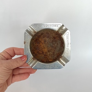 May include: Vintage metal ashtray with a square shape and a circular, rusty interior. The ashtray has the words "PIPE WRENCHES" and "THREADERS-CUTTERS" embossed on the sides. The brand name "RIDGID MARK" is also visible.