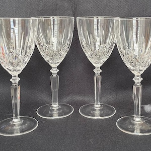 May include: Four clear crystal wine glasses with intricate etched patterns. Each glass features a rounded bowl, a long, rectangular stem, and a circular base. The glasses are arranged on a dark surface.