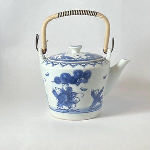 May include: A white porcelain teapot with a blue and white design featuring figures and a tree. The teapot has a woven handle with a black and white pattern.