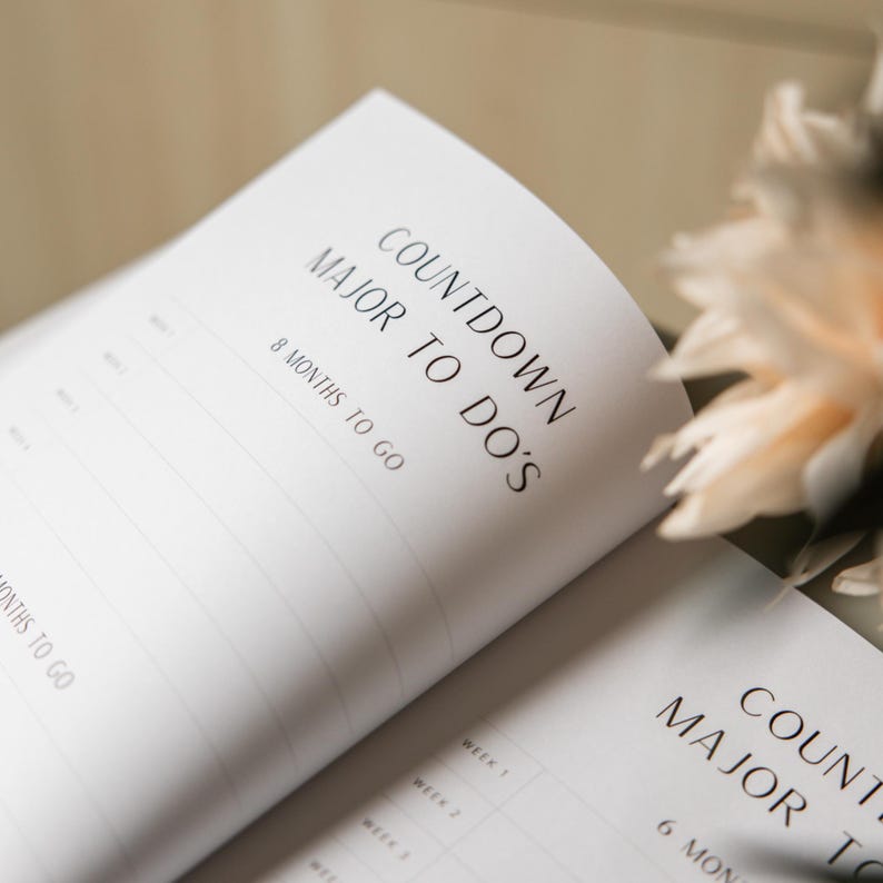 May include: Open planner with the text "COUNTDOWN MAJOR TO DO'S" and "8 MONTHS TO GO" in black font. The pages are white with horizontal lines for notes. A soft, blurred floral arrangement is in the background.