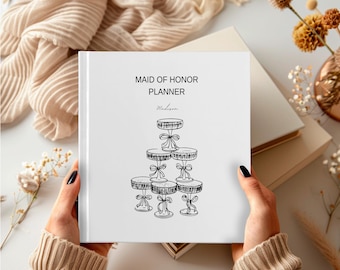 Puede incluir: Una agenda blanca titulada "MAID OF HONOR PLANNER" con una ilustración negra de soportes para pasteles escalonados. La agenda está sostenida por manos que llevan un suéter beige. Tela beige y un jarrón con flores secas en el fondo.