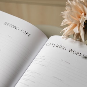 May include: An open notebook with the headings "WEDDING CAKE" and "CATERING WORKSHOP" printed in black. The pages are lined and blank, with space for notes. A bouquet of light-colored flowers is in the background.