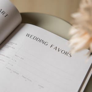 May include: Open notebook with the headline "WEDDING FAVORS" in a minimalist design. The pages are white with black text and lines for writing. The notebook is open on a light-colored surface.