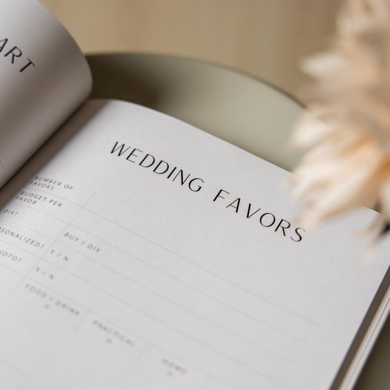 May include: Open notebook with the headline "WEDDING FAVORS" in a minimalist design. The pages are white with black text and lines for writing. The notebook is open on a light-colored surface.