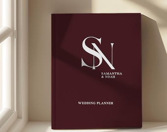 May include: A burgundy wedding planner with the initials "SN" in silver. The names "Samantha & Noah" are printed below the initials, and the words "WEDDING PLANNER" are at the bottom. The planner is on a white surface.