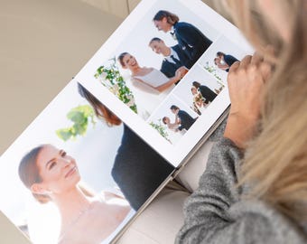 May include: A white photo album open to a spread of wedding photos. The album displays multiple images of a bride and groom, capturing moments from their wedding day. The photos are in color and show the couple smiling and embracing.