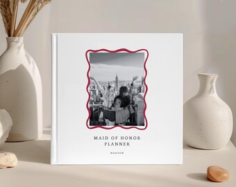 Puede incluir: Un planificador blanco con un borde ondulado burdeos alrededor de una foto en blanco y negro de dos personas en una azotea. El texto "MAID OF HONOR PLANNER" y "MADISON" está impreso debajo.
