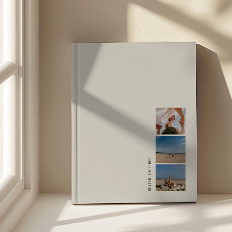 May include: A pale beige photo album featuring three small photographs and the text "BETTER TOGETHER" printed vertically. The photographs depict people on a beach. The album is positioned on a white surface near a window.