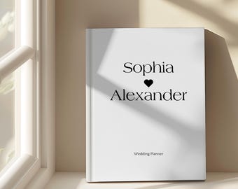 May include: A white wedding planner book with the names "Sophia" and "Alexander" in black script, separated by a black heart. The words "Wedding Planner" are printed below. The book is displayed near a window.