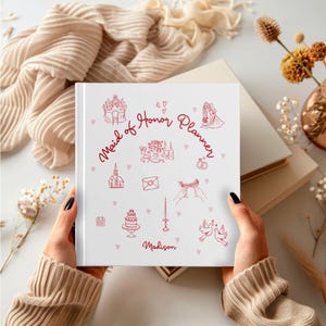 May include: A white "Maid of Honor Planner" book with red illustrations of wedding-related icons. The book is held by a person wearing a beige jumper. The text "Maid of Honor Planner" is written in red script, with the name "Madison" below.