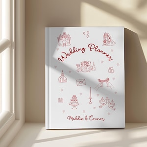 May include: A white wedding planner book featuring red illustrations of wedding symbols like a church, cake, and rings. The words "Wedding Planner" and the names "Maddie & Connor" are written in red script.