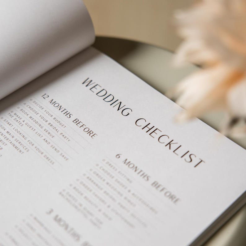 May include: Open wedding checklist book with the title "WEDDING CHECKLIST" in elegant black font. The checklist is divided into sections like "12 MONTHS BEFORE" and "6 MONTHS BEFORE", with bullet points for tasks like "DECIDE YOUR BUDGET" and "CHOOSE YOUR BRIDAL PARTY".