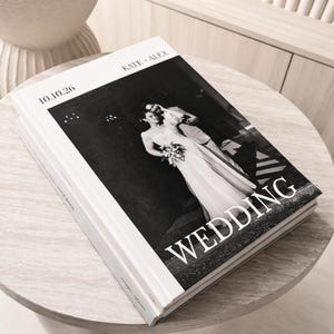 May include: A stack of white wedding photo books with the names "KATE + ALEX" and the date "10.10.26" printed on the cover. The book's cover features a black and white photo of a bride and groom with the word "WEDDING" in large white letters.