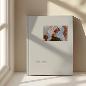 May include: A cream-coloured photo album with the words "BETTER TOGETHER" printed on the front. The album features a small, rectangular photo of two people looking up at each other. The background is a light beige.