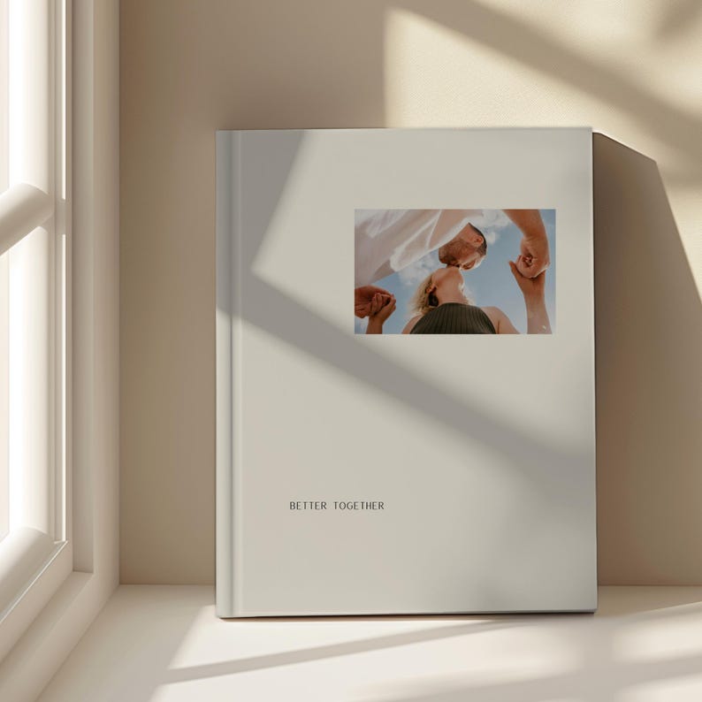 May include: A cream-coloured photo album with the words "BETTER TOGETHER" printed on the front. The album features a small, rectangular photo of two people looking up at each other. The background is a light beige.