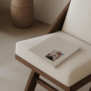 May include: A book with a beige cover and a small image rests on a white upholstered chair. The chair has a dark wooden frame. The book's title is visible. A decorative vase is in the background.