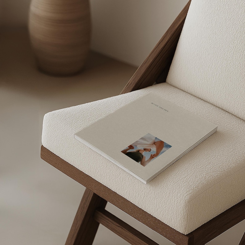 May include: A book with a beige cover and a small image rests on a white upholstered chair. The chair has a dark wooden frame. The book's title is visible. A decorative vase is in the background.