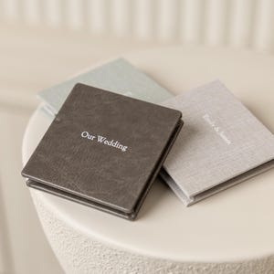 May include: Three small, square wedding photo albums in shades of grey and brown are displayed. The front album is dark grey with the words "Our Wedding" in white. The other two albums are light grey and green, with the names "Emily & James" on one.