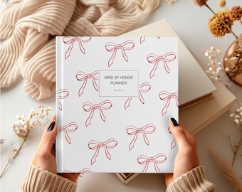 Puede incluir: Una "Maid of Honor Planner" blanca con un patrón de lazo rojo. La agenda la sostiene una persona que lleva un suéter beige. El fondo incluye una bufanda beige, un jarrón con flores secas y otros artículos de papelería.