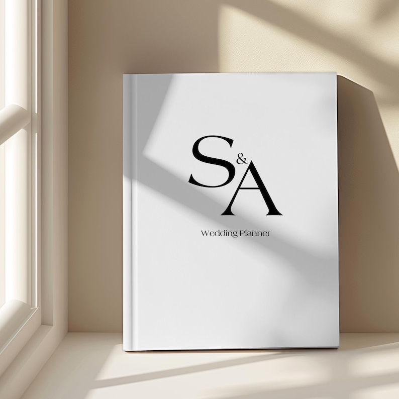 May include: A white wedding planner book with the initials "S&A" in black, and the words "Wedding Planner" below. The book is standing upright on a white surface, next to a window.