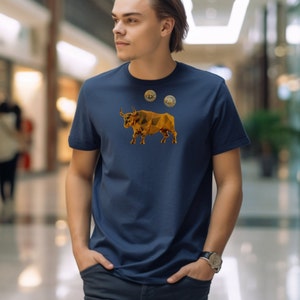Puede incluir: Una camiseta azul marino con un diseño de toro geométrico dorado y dos monedas de oro en la parte delantera.