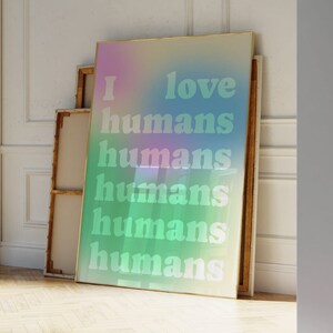 May include: A framed art print with a pastel gradient background of pink, blue, and green. The words "I love humans" are printed in a light green font. The print leans against a wall in a room with a wooden floor.