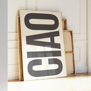 May include: A large framed artwork with the word "CIAO" in bold, black letters against a cream background. The artwork leans against a white wall, with other framed pieces. The room has a hardwood floor and an open doorway.