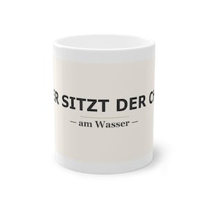 Könnte beinhalten: Weißer Keramikbecher mit dem Text "ER SITZT DER C - am Wasser -" in Schwarz gedruckt. Der Becher hat ein cremefarbenes Band in der Mitte. Der Becher ist zylindrisch und hat einen weißen Henkel.