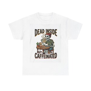 Könnte beinhalten: Weißes T-Shirt mit einer Grafik eines Skeletts mit Mütze, das Kaffee trinkt, und dem Text "DEAD INSIDE BUT CAFFEINATED". Das Skelett sitzt an einem Tisch mit einer Kaffeetasse und einem kleinen Kaktus.