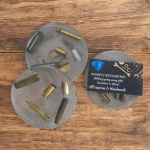 May include: Three clear resin coasters with embedded brass and silver bullet casings. One coaster has a business card that reads "HUGHEY'S WOODWORKS Military going away gifts Furniture & More! All custom & Handmade."