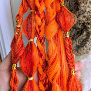 Puede incluir: Extensiones de pelo sintético naranja y rojo con cuentas doradas, trenzadas y atadas juntas con gomas elásticas.
