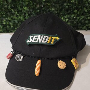 Peut inclure: Casquette de baseball noire avec le texte "SEND IT" en vert et blanc. La casquette a une chaîne avec des breloques représentant une canette de soda, une gemme, une baguette, un biscuit et un paquet de chips.