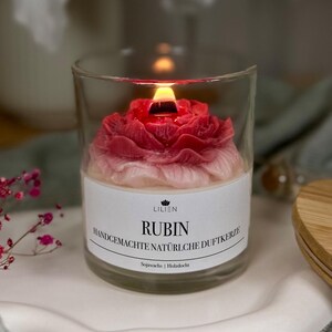 Könnte beinhalten: Eine brennende, rubinrote Kerze in Rosenform, umhüllt von einem klaren Glas. Die Kerze hat ein weißes Etikett mit dem Wort "RUBIN" und anderem Text. Die Kerze ist handgefertigt und aus Sojawachs.