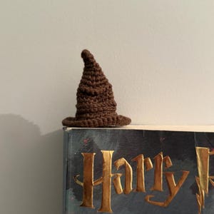 Può includere: Un piccolo cappello da mago marrone all'uncinetto, con una corona a punta e un'ampia tesa. Il cappello è appoggiato su un libro con lettere dorate che dicono "Harry". Il cappello è fatto di filato strutturato.