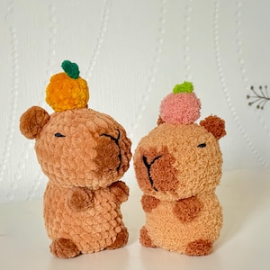 Peut inclure: Deux capybaras au crochet faits à la main, dans des tons de marron et de beige. Chaque capybara porte un petit pompon sur la tête, l'un orange avec une feuille verte et l'autre rose avec une feuille verte. Les capybaras ont des traits du visage brodés en noir.