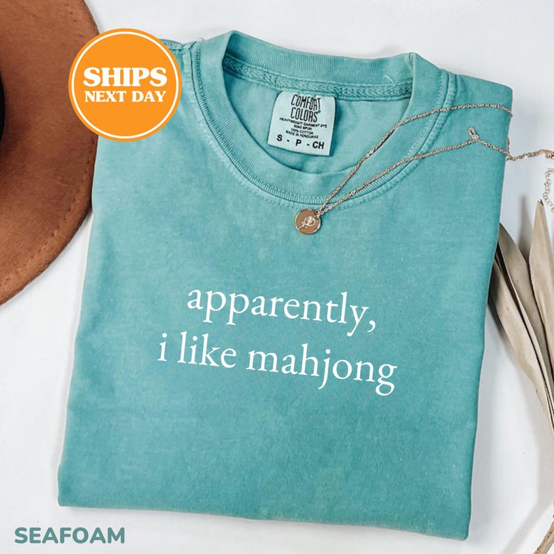May include: Seafoam green t-shirt with the text "apparently, i like mahjong" in white. The shirt has a crew neck and short sleeves. A gold necklace with a pendant is draped across the shirt.