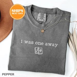 May include: A charcoal gray short-sleeve t-shirt with a crew neckline. The front features the white text "i was one away" above a graphic of two mahjong tiles. A thin silver chain necklace with a small circular pendant rests on the collar.