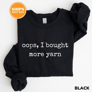 A black t-shirt with white text printed on it, which says "oops, I bought more yarn".