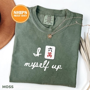 Puede incluir: Una camiseta de manga corta color verde musgo con el texto blanco "I [ficha de mahjong] myself up" y un gráfico de una ficha de mahjong que muestra el carácter chino para 60.000 en negro y rojo. La camiseta tiene cuello redondo y etiqueta Comfort Colors.