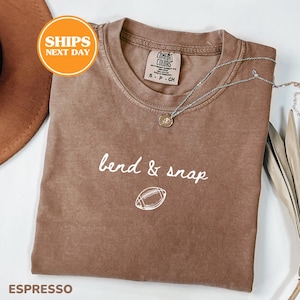 May include: A brown short-sleeved t-shirt with a crew neckline featuring the white text bend & snap and a white graphic of a football. The shirt has a Comfort Colors brand label on the inside collar and is displayed with a silver-toned necklace.