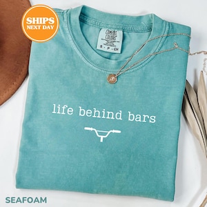 A turquoise t-shirt with the text "life behind bars" printed on it, along with a bicycle graphic. The t-shirt is folded and placed on a white surface, with a brown hat and some leaves visible in the background.